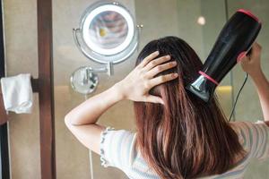 jovem mulher usando secador de cabelo perto do espelho em casa ou hotel. penteados e conceitos de estilo de vida foto