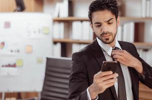 retrato de empresário confiante usando smartphone na sala de trabalho. bonito empresário árabe de terno preto e olhando o celular no escritório. foto