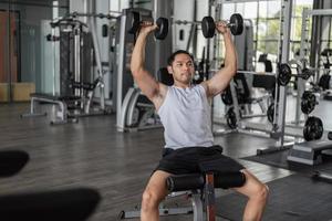 homem asiático exercita em fitness. jovem saudável em roupas esportivas fazendo exercícios de ombros no ginásio. foto