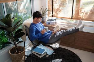 homem asiático bonito usando óculos e sorrindo enquanto lê o livro em casa sala de estar, literatura e conceito de lazer