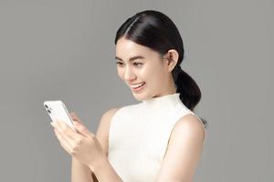 mulher asiática confiante usando smartphone e recebendo boas notícias da mensagem no aplicativo de bate-papo móvel isolado em fundo cinza. retrato de uma linda garota no estúdio.