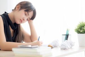 linda mulher asiática cansada e estressada com a escrita sobrecarregada na mesa, garota com ideia não preocupada com caderno e papel amassado no escritório, freelance e conceito de negócios. foto