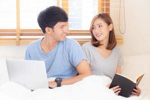 jovem casal asiático com homem usando computador portátil e pesquisando internet e mulher lendo notebook na cama no quarto com feliz e relaxe, comunicação e conceito de negócios. foto