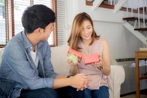 jovem casal asiático comemora aniversário juntos, homem asiático dando presente de caixa de presente para mulher para surpresa na sala de estar, feminino se sentindo feliz e animado aniversário, conceito de feriado dos namorados. foto