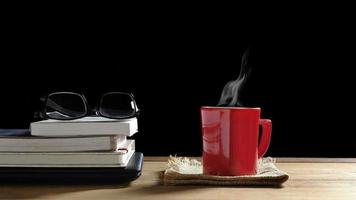 café quente em xícara vermelha com vapor e óculos em livros empilhados com laptop na mesa de madeira em fundo preto, conceito de pausa para o café foto