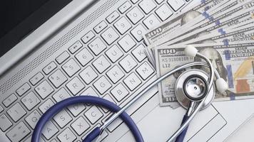 postura plana de estetoscópio com notas de dólar no teclado do laptop, saúde do mercado financeiro e conceito de despesas médicas foto
