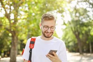 retrato de jovem barbudo bonito com fones de ouvido, olhando para o telefone, sendo positivo e satisfeito, de pé sobre as árvores verdes em dia ensolarado foto