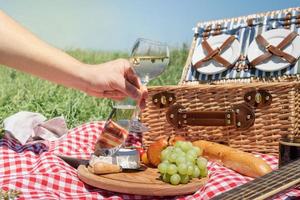closeup de cesta de piquenique com bebidas e comida na grama. mão masculina segurando o copo de vinho foto