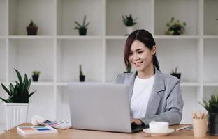 alegre e atraente jovem designer gráfico feminino sorrindo e trabalhando no computador portátil em sua mesa no escritório moderno foto