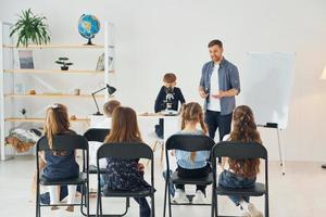 olhando para o menino com microscópio que está em cima da mesa. grupo de alunos de crianças em sala de aula na escola com professor foto