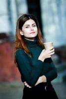 jovem elegante bebendo chá em uma rua da cidade. menina europeia hipster com vidro de papel com leite. linda jovem com café na rua da cidade. pausa para o café. café para viagem. foto