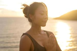 jovem mulher asiática em roupas esportivas fazendo ioga na rocha à beira-mar durante o conceito de pôr do sol, saúde e meditação