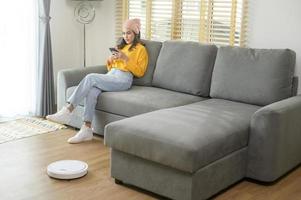 jovem mulher feliz relaxando e usando smartphone na sala de estar enquanto aspirador de pó robótico trabalhando foto