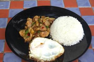 manjericão, frango, ovo frito, comida tailandesa o estilo da casa não é sofisticado em uma placa de cerâmica preta colocada em uma mesa de concreto xadrez. pronto para servir e entrega foto