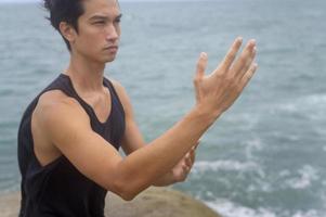 jovem em roupas esportivas fazendo arte marcial, qigong, tai chi na rocha no conceito de beira-mar, saúde e meditação foto