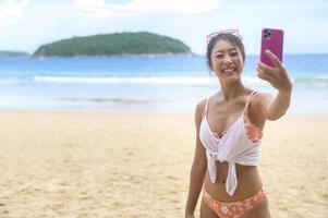 jovem mulher bonita de biquíni ouvindo música e usando smartphone na praia, verão, férias, feriados, conceito de estilos de vida. foto