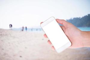 mão de mulher mostrando um telefone inteligente em branco com telas isoladas no fundo da praia foto