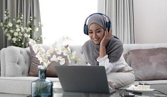 lista de reprodução para retrato de relaxamento de uma mulher muçulmana calma em hijab ouvindo música com seu laptop e fones de ouvido sem fio em casa. uma mulher islâmica sorridente senta-se com os olhos fechados foto