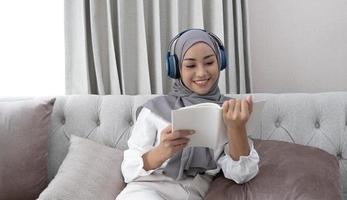 encantadora jovem muçulmana asiática usando hijab e fones de ouvido, ouvindo música e lendo um livro na sala de estar. foto