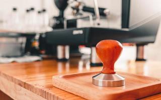 close-up de um calcador de café de ferramenta de barista com cabo de madeira colocado em um balcão de bar de madeira com uma máquina de café em uma cafeteria. foto