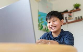 sorridente jovem asiático conversando com amigos ou um estudante estudando a internet em redes sociais sentado na frente do laptop em casa. bate-papo nas redes sociais e videoconferência. foto