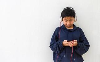 menino asiático bonito com pele branca em um capuz posando ouvindo a música em um telefone celular e fones de ouvido isolados sobre fundo branco. espaço para retrato e cópia foto