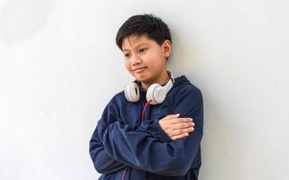 menino asiático bonito em um capuz posando em pé sorrindo com os braços cruzados em um gesto feliz e confiante usando fones de ouvido isolados no fundo branco retrato e estilo de vida da criança foto