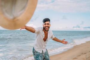homem latino-americano sorrindo e jogando seu chapéu olhando para a câmera na praia em um lindo dia de verão foto