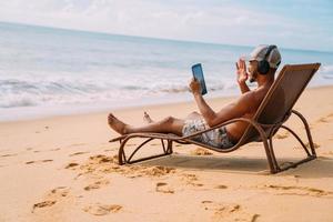 homem fazendo uma chamada de vídeo nas férias de verão. homem latino-americano sentado na cadeira de praia com fones de ouvido e um tablet foto