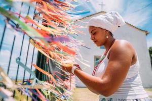 mulher brasileira vestida com o traje tradicional baiano da religião umbandista fazendo uma oração. foco em fitas coloridas de promessas religiosas em devoção ao senhor do bonfim na bahia, brasil foto