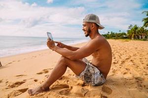 férias de verão, tecnologia e conceito de internet. homem latino-americano sentado na areia, tomando sol na praia com um tablet foto