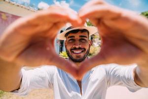 turista fazendo selfie no centro histórico de porto seguro. homem latino-americano de chapéu, fazendo um coração com as mãos, sorrindo para a câmera foto