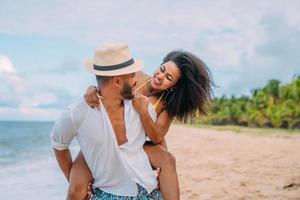 jovem casal em férias de verão na praia, homem sorridente feliz carrega mulher de volta à beira-mar foto