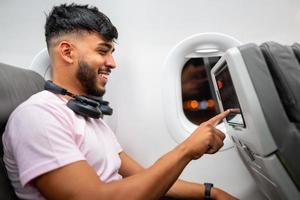 passageiro no avião tocando a tela de entretenimento lcd. homem latino-americano na cabine do avião, usando dispositivo inteligente, com fones de ouvido. foto