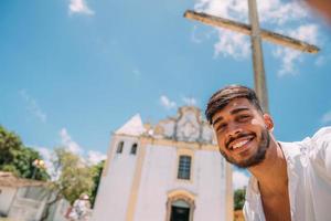 amigável jovem latino-americano sorrindo para a câmera. turista tirando uma selfie com a igreja de nossa senhora da ajuda ao fundo foto