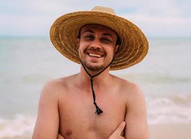 jovem latino-americano na praia, com um chapéu e braços cruzados. homem sorridente, olhando para a câmera foto