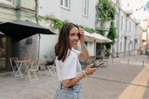 senhora muito adorável com cabelo castanho virar para a câmera e posar para a câmera enquanto caminhava na rua e usando smartphone foto