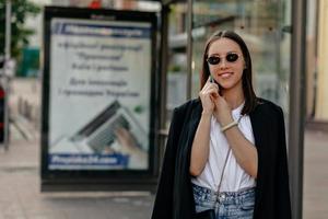 atraente senhora bonita com cabelos escuros vestindo jaqueta preta uma camisa branca em óculos de sol está falando ao telefone e andando na cidade com emoções felizes foto