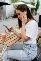 encantadora senhora adorável de camiseta branca e jeans tocando fones de ouvido e ouve música ao ar livre no terraço de verão. mulher bonita lin fones de ouvido sem fio no terraço de verão foto