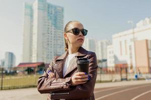 senhora elegante independente com cabelos escuros usando óculos escuros e jaqueta bordô está olhando de lado enquanto caminhava na cidade com café à luz do sol foto