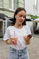 mulher adorável espetacular com um lindo sorriso segurando smartphone e smoothie de verão e olhando para smartphone. foto ao ar livre de senhora encantadora com gadget