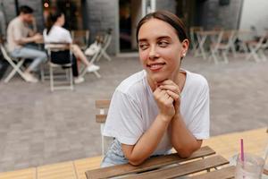 linda mulher com sorriso feliz, vestindo camiseta branca está de mãos dadas perto do rosto e olhando de lado com um sorriso feliz, sentado na cafeteria da rua e esperando os amigos foto