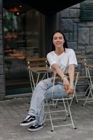 retrato completo de encantadora senhora bonita em jeans e camiseta branca está descansando do lado de fora na cafeteria da rua e sorrindo para a câmera com emoções felizes foto