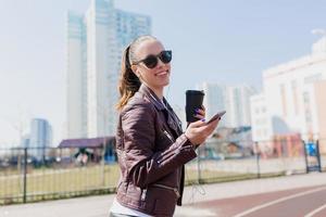 retrato outsdie de encantadora mulher bonita sorridente com cabelo escuro vestido de jaqueta de couro e óculos escuros ouvindo música em fones de ouvido na luz solar foto