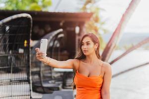 mulher jovem de raça mista tomando uma selfie em um telefone celular com vestido laranja de verão. foto