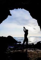 silhueta de uma mulher levantando as mãos para ioga ou posando para abrir a mente para coisas bonitas. foto de uma mulher praticando ioga em pé em uma caverna com vista para o mar ao nascer ou pôr do sol.