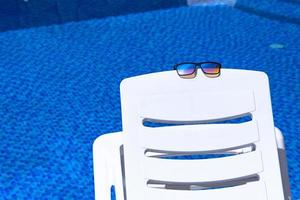 óculos de sol estão em uma espreguiçadeira branca contra o pano de fundo de uma piscina com água azul. espaço de cópia foto