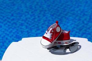 óculos de sol e sapatos de ginástica infantil vermelhos estão em uma espreguiçadeira branca contra o pano de fundo de uma piscina com água azul. espaço de cópia foto