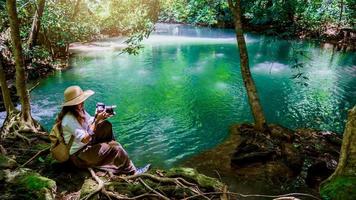 menina com uma mochila amarela está andando na natureza de viagens. viajante elegante hipster andando na floresta tropical ensolarada. belos riachos esmeralda e árvores verdes na floresta tropical. viajando tirando fotos