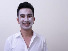 jovem bonito asiático aplicando creme no rosto com carinha sorridente, conceito de cuidados com a pele foto